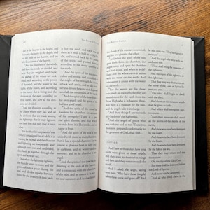 Puede incluir: Un libro de tapa dura abierto con p&aacute;ginas blancas y texto impreso en negro. La p&aacute;gina izquierda se titula The Book of Enoch y contiene varios p&aacute;rrafos de texto. La p&aacute;gina derecha contin&uacute;a el texto e incluye el encabezado Chapter 61.