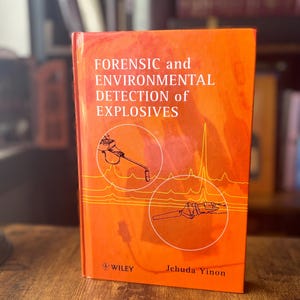 May include: A hardcover book titled Forensic and Environmental Detection of Explosives by Jehuda Yinon, published by Wiley. The cover is bright orange with white text and features two circular line drawings of scientific equipment and data graphs.