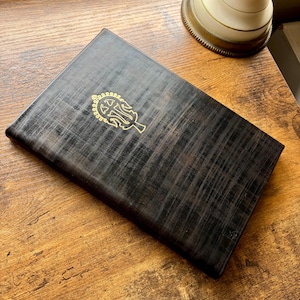 Puede incluir: Una caja rectangular de madera en tonos oscuros con un patr&oacute;n de veta entrecruzada visible. La tapa presenta un emblema met&aacute;lico dorado centrado que representa un s&iacute;mbolo estilizado y ornamentado en forma de &aacute;rbol con ramas sim&eacute;tricas.