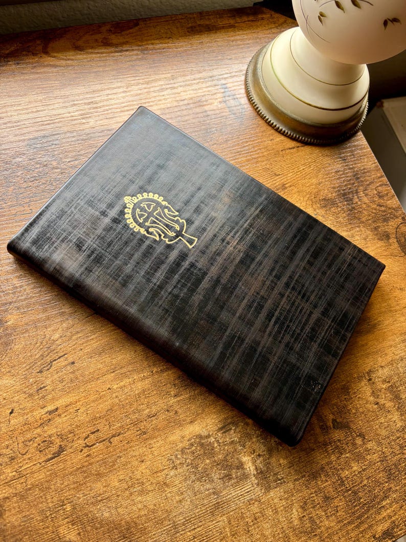 Puede incluir: Una caja rectangular de madera en tonos oscuros con un patr&oacute;n de veta entrecruzada visible. La tapa presenta un emblema met&aacute;lico dorado centrado que representa un s&iacute;mbolo estilizado y ornamentado en forma de &aacute;rbol con ramas sim&eacute;tricas.