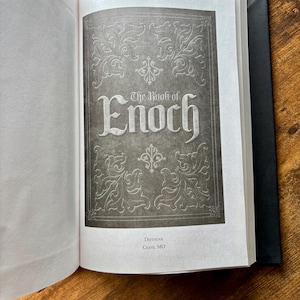 Puede incluir: Un libro de tapa dura abierto que muestra una p&aacute;gina de t&iacute;tulo con el texto The Book of Enoch en una fuente estilizada. La p&aacute;gina presenta un fondo gris oscuro con bordes florales decorativos blancos y el texto Defender Crane, MO abajo.