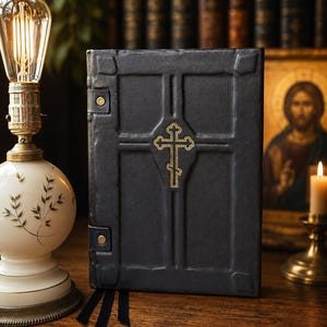 May include: A black leather-bound book featuring a raised cross design on the front cover with a gold-colored embroidered Orthodox cross in the center. The cover includes two side straps with gold-colored metal studs and three black ribbon bookmarks.