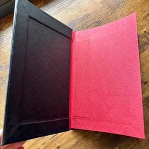 Puede incluir: Una carpeta o portafolio rectangular con un marco interior negro en el lado izquierdo y un panel de papel rojo s&oacute;lido en el lado derecho. El objeto est&aacute; abierto, revelando un esquema de color contrastante con una textura mate en ambas superficies.