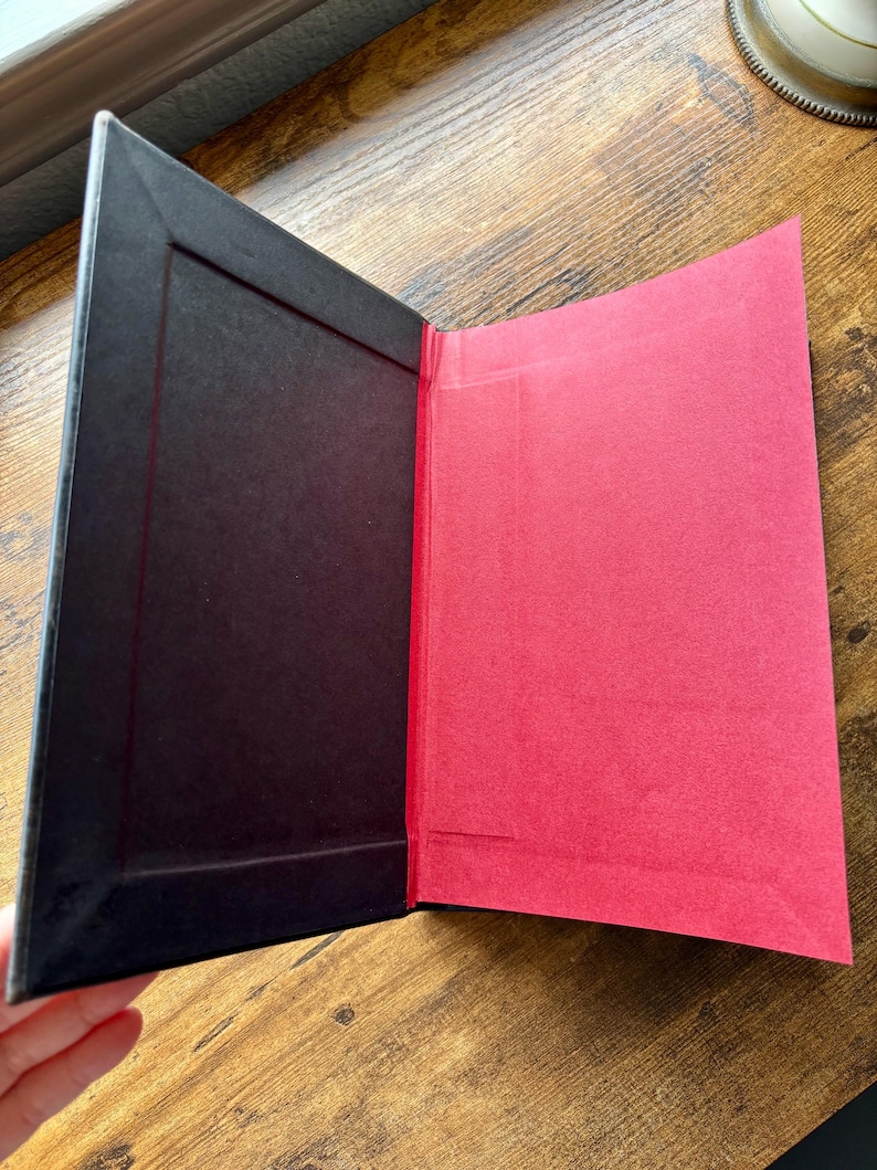 Puede incluir: Una carpeta o portafolio rectangular con un marco interior negro en el lado izquierdo y un panel de papel rojo s&oacute;lido en el lado derecho. El objeto est&aacute; abierto, revelando un esquema de color contrastante con una textura mate en ambas superficies.