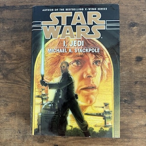 May include: A hardcover book titled "Star Wars I, Jedi" by Michael A. Stackpole. The cover features a depiction of a Jedi with a lightsaber, R2-D2, and a portrait of a man. The Star Wars logo is prominently displayed at the top.