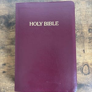 May include: A burgundy-colored Holy Bible with gold lettering. The book is closed and has a textured cover. The title "HOLY BIBLE" is prominently displayed on the front cover. The book is resting on a wooden surface.