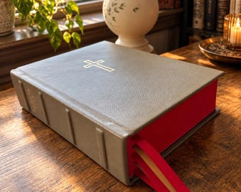 May include: A thick, gray, leather-bound book featuring a gold-stamped cross on the front cover. The book has a textured surface, raised spine bands, and bright red page edges with three attached ribbon bookmarks in red and cream colors.