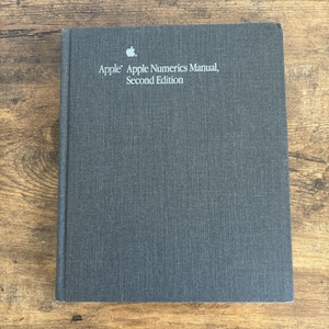 May include: A dark gray book titled "Apple Apple Numerics Manual, Second Edition" with the Apple logo. The book has a textured cover and is lying on a wooden surface. The book is a manual.