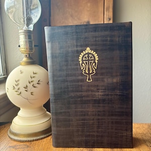 Puede incluir: Un libro de tapa dura de color marr&oacute;n oscuro con textura y un emblema dorado de una cruz en la portada. El libro est&aacute; colocado junto a una l&aacute;mpara de mesa con una base de cristal blanca y redondeada decorada con hojas verdes y una bombilla.