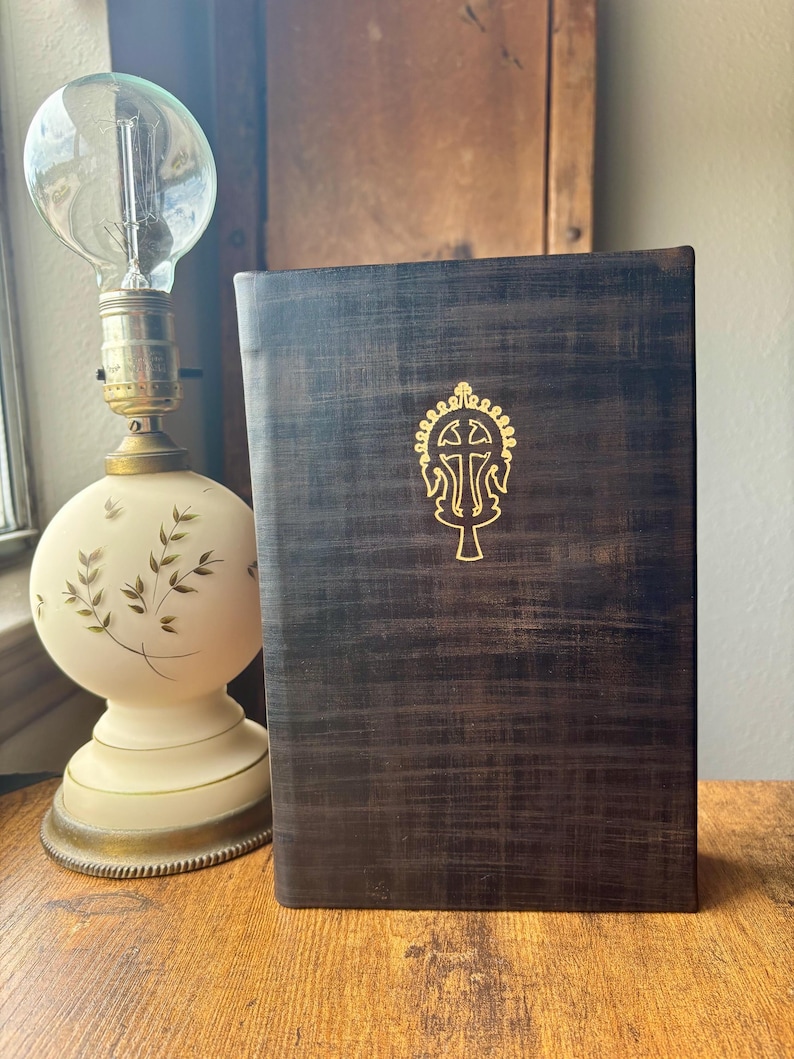 Puede incluir: Un libro de tapa dura de color marr&oacute;n oscuro con textura y un emblema dorado de una cruz en la portada. El libro est&aacute; colocado junto a una l&aacute;mpara de mesa con una base de cristal blanca y redondeada decorada con hojas verdes y una bombilla.