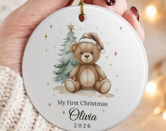 May include: A round white ceramic Christmas ornament featuring a brown teddy bear wearing a Santa hat next to a green pine tree. The text reads My First Christmas, the name Olivia, and the year 2026. It hangs from a gold string.