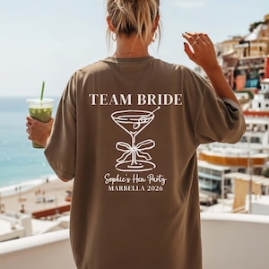 Op de afbeelding: Een espresso-kleurig t-shirt met witte tekst en een martini-glasafbeelding op de achterkant. De tekst luidt TEAM BRIDE, Sophie's Hen Party en MARBELLA 2026. Het shirt heeft korte mouwen en een ronde hals, gezien vanaf de achterkant.