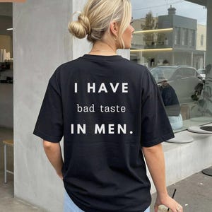 Op de afbeelding: Zwart T-shirt met korte mouwen met de witte tekst "I HAVE bad taste IN MEN.". Het shirt wordt gedragen door een persoon met blond haar, die buiten een gebouw staat. Het woord "BLACK" staat in de linkerbovenhoek.