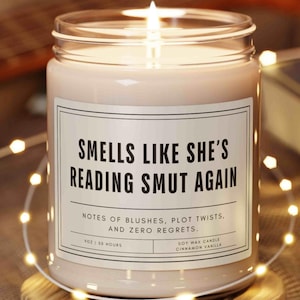 May include: A lit soy wax candle in a clear glass jar with a white label reading "SMELLS LIKE SHE'S READING SMUT AGAIN" and "NOTES OF BLUSHES, PLOT TWISTS, AND ZERO REGRETS." The label also states "9OZ | 50 HOURS" and "CINNAMON VANILLA."