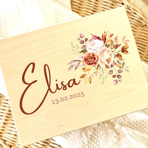 Puede incluir: Una caja de madera de color claro con el nombre "Elisa" en una fuente marrón y la fecha "13.02.2025". La caja está decorada con un arreglo floral en tonos melocotón, burdeos y dorado. La caja descansa sobre una manta blanca.