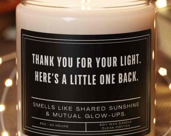 Może przedstawiać: Płonąca świeca z wosku sojowego w przezroczystym szklanym słoiku z czarną etykietą o treści: Thank you for your light. Here's a little one back. Smells like shared sunshine & mutual glow-ups. 9oz 50 hours soy wax candle clean cotton. Świeca stoi na drewnianej podstawie.