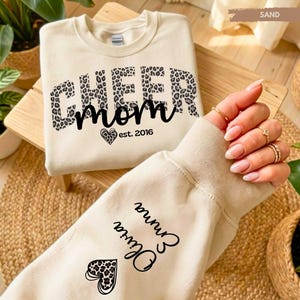 Könnte beinhalten: Ein cremefarbenes Sweatshirt mit den Worten "CHEER mom" in Schwarz und Leopardenmuster. Der Ärmel zeigt die Namen "Olivia Emma" in schwarzer Schrift. Ein Herz mit Leopardenmuster ist ebenfalls sichtbar.