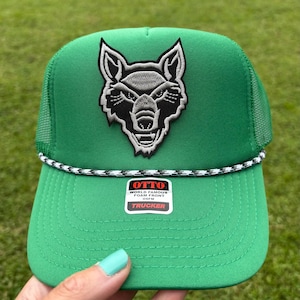 May include: Green trucker hat with a wolf patch. The hat features a mesh back, a curved brim, and a black and white rope detail. The patch is a detailed embroidered wolf design. The hat has a tag that says "OTTO WORLD FAMOUS FOAM FRONT TRUCKER".