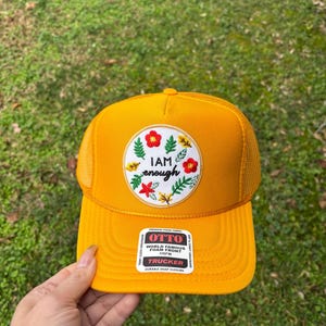Op de afbeelding: Mosterdgele truckerpet met een witte patch met de tekst "I AM enough" omringd door rode bloemen en groene bladeren. De pet heeft een mesh achterkant en een druksluiting.