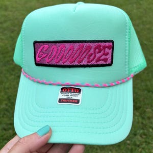 May include: A mint green trucker hat with a pink and black embroidered patch that reads "GOODSE". The hat has a mesh back and a pink and blue rope detail along the brim. The hat has a "World Famous Foam Front" sticker.