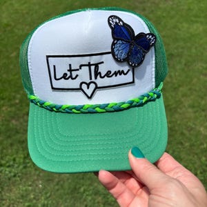 May include: A green and white trucker hat featuring a white front panel with a black rectangular patch containing the text Let Them and a heart symbol. A blue butterfly patch is attached above the text. A braided green and blue cord trim.