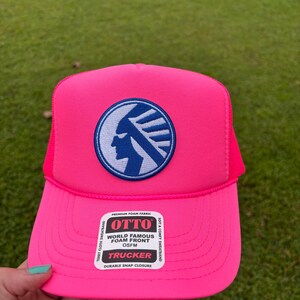 Peut inclure: Casquette de camionneur rose vif avec un écusson circulaire bleu et blanc représentant un profil amérindien. Le chapeau a un devant en mousse et un dos en filet. L'étiquette indique "OTTO WORLD FAMOUS FOAM FRONT TRUCKER".