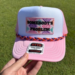 May include: Pink and white trucker hat with the text "SOMEBODY'S PROBLEM" on the front. The hat has a pink brim and mesh back, with a braided rope detail. The hat also has a "OTTO" label.