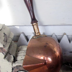 May include: A vintage copper coal scuttle with a dark wooden handle and brass collar. The scuttle has a rounded body and a small spout. The handle is attached to the scuttle with a brass fitting. The scuttle is likely used for carrying coal or other solid fuels.