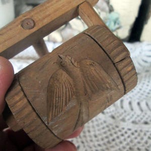 May include: A hand-carved wooden object with a bird design. The cylindrical piece is brown with a detailed carving of a bird with outstretched wings. The wood has a natural grain and shows signs of age.