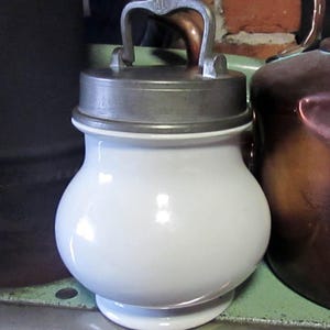 May include: A white, bulbous ceramic jar with a metal lid and handle. The lid is attached to the jar with a hinge. The handle has the letters "AL" on it. The jar is sitting on a green surface.