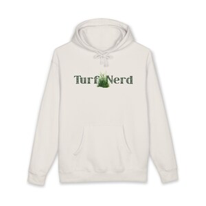 Puede incluir: Una sudadera con capucha blanca con un bolsillo delantero y un cordón. El texto "Turf Nerd" está impreso en el pecho en una fuente verde texturizada, con un pequeño parche de hierba que reemplaza la letra "u".
