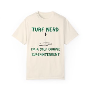May include: A beige t-shirt with the text "TURF NERD" and "I'M A GOLF COURSE SUPERINTENDENT" in green text. A green golf flag and a golf hole are pictured on the shirt.