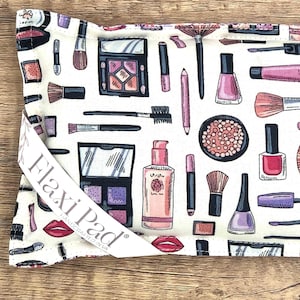 May include: White cosmetic bag with a repeating pattern of makeup items in pink, purple, and black. The design includes lipsticks, brushes, eyeshadow palettes, and nail polish. The bag has a white strap that reads "Flaxi Pad".