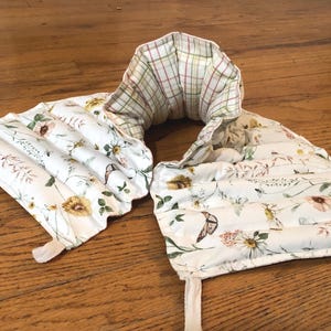 May include: A floral-patterned neck wrap with a cream background, featuring yellow, pink, and brown flowers, green leaves, and butterflies. The wrap has a plaid lining and a fabric tie. The product is designed to be placed around the neck.