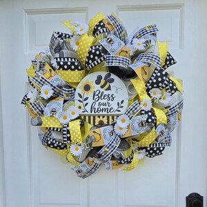 May include: A decorative wreath featuring yellow and black mesh ribbons, polka dot and buffalo check patterns, white daisy accents, and bee illustrations. A central circular sign displays the text Bless our home with a bee and sunflower graphic.
