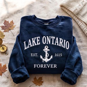 May include: A navy blue crewneck sweatshirt featuring white embroidered text that reads LAKE ONTARIO, EST. 1615, and FOREVER, with a central white anchor graphic. The garment has long sleeves with ribbed cuffs and a ribbed neckline.