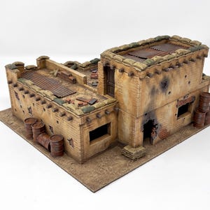 May include: A miniature two-storey desert building model featuring tan textured walls with dark weathering, bullet holes, and scorch marks. The structure includes sandbag fortifications on the roof, corrugated metal roofing, and brown barrels.