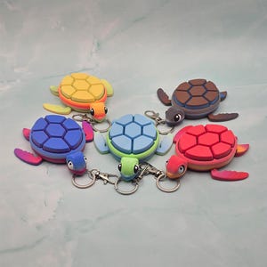 May include: A selection of turtle-shaped keyrings in various colours. The keyrings are in shades of yellow, blue, green, red, and brown. Each turtle has a segmented shell design and is attached to a silver keyring.