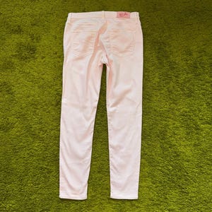 May include: A pair of light pink denim pants featuring a slim-fit silhouette, two back pockets, and a rectangular pink brand label stitched onto the waistband. The pants have a classic five-pocket construction with visible stitching and a button closure.