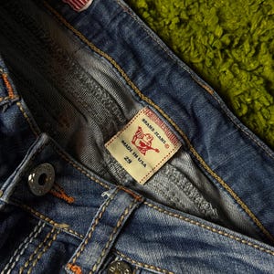 May include: Close-up of blue denim jeans with orange and white stitching. Features a small American flag patch and a label that reads "TRUE RELIGION BRAND JEANS" and "MADE IN USA 29". The jeans have silver metal buttons.