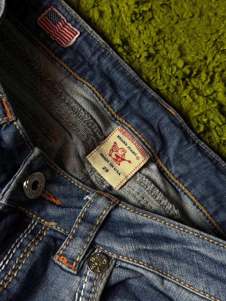 May include: Close-up of blue denim jeans with orange and white stitching. Features a small American flag patch and a label that reads "TRUE RELIGION BRAND JEANS" and "MADE IN USA 29". The jeans have silver metal buttons.