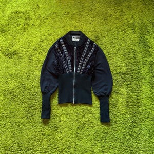 May include: Black bomber jacket with a velvet panel on the chest, featuring silver embellishments and a zipper closure. The jacket has long sleeves with fitted cuffs and a ribbed waistband. The background is a bright green carpet.