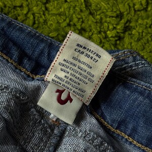 May include: Close-up of a pair of blue denim jeans with a white tag. The tag displays the RN and CA numbers, along with washing instructions and the fabric content, 100% cotton. The True Religion logo is visible.