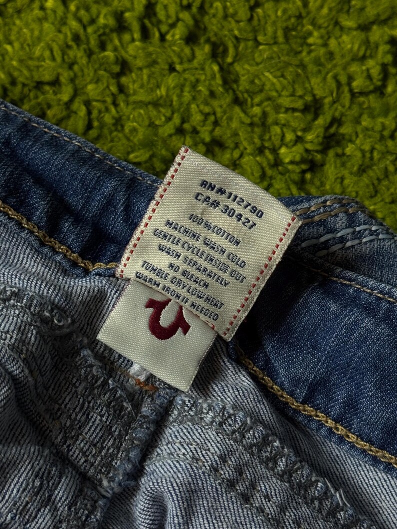 May include: Close-up of a pair of blue denim jeans with a white tag. The tag displays the RN and CA numbers, along with washing instructions and the fabric content, 100% cotton. The True Religion logo is visible.