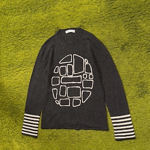 May include: A dark gray sweater with a white geometric design on the front. The long sleeves have white and black striped cuffs. The sweater is displayed on a green surface.