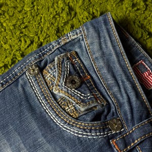 May include: Close-up of blue denim jeans with decorative stitching and metal button details. The jeans feature a pocket with a unique design and an American flag patch. The stitching is white and orange, adding contrast to the denim.