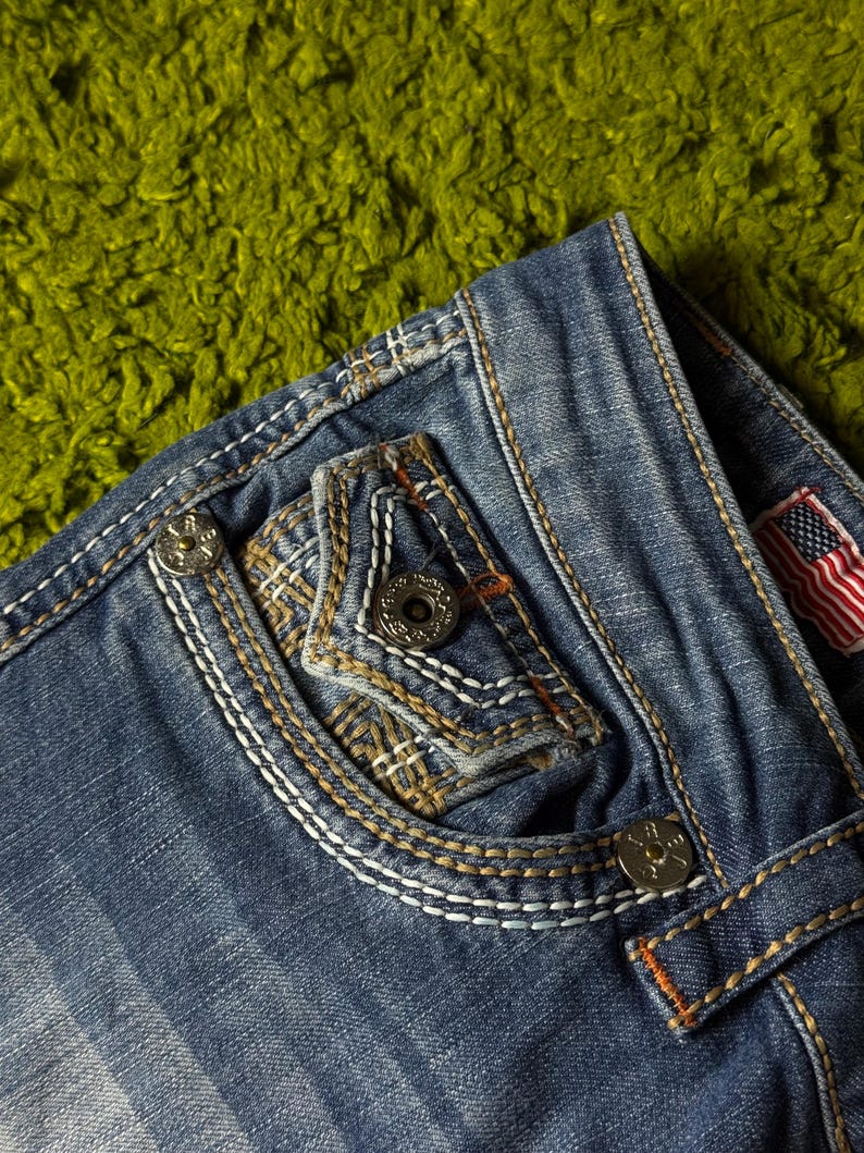 May include: Close-up of blue denim jeans with decorative stitching and metal button details. The jeans feature a pocket with a unique design and an American flag patch. The stitching is white and orange, adding contrast to the denim.