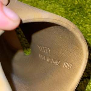 K&ouml;nnte beinhalten: Nahaufnahme des Inneren einer beigefarbenen Sandale. Die Innensohle der Sandale tr&auml;gt den Markennamen "YEEZY", den Text "MADE IN CHINA" und die Gr&ouml;&szlig;e "6US" in Schwarz.