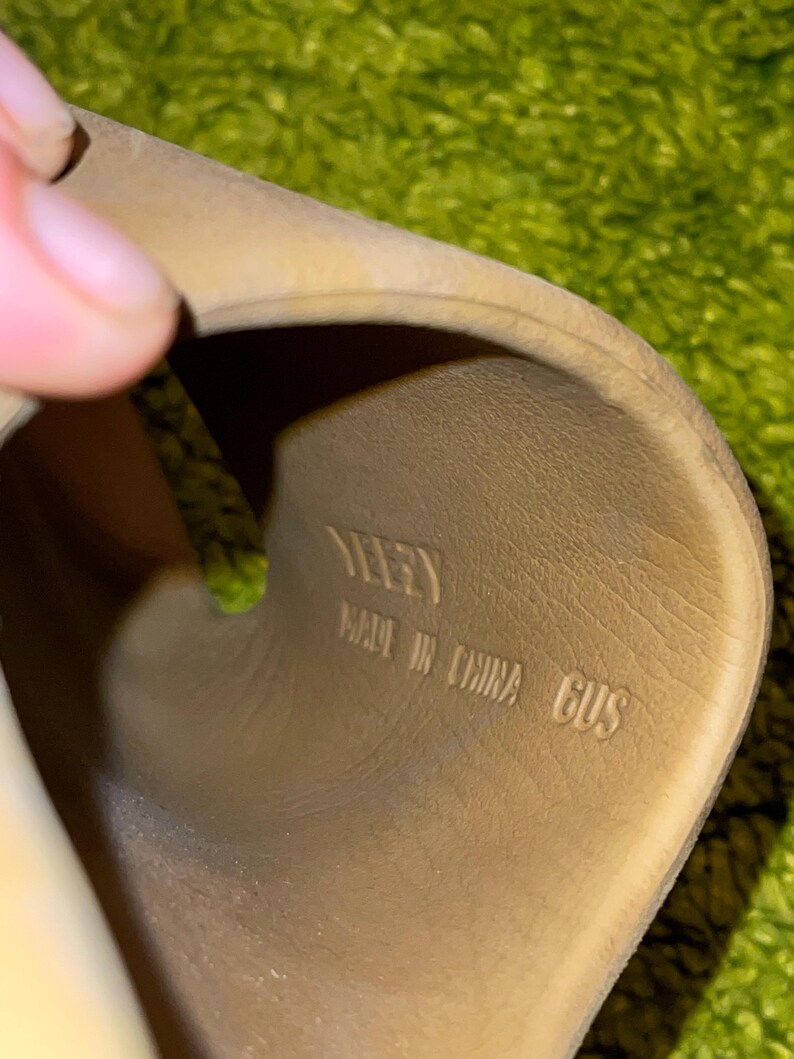 K&ouml;nnte beinhalten: Nahaufnahme des Inneren einer beigefarbenen Sandale. Die Innensohle der Sandale tr&auml;gt den Markennamen "YEEZY", den Text "MADE IN CHINA" und die Gr&ouml;&szlig;e "6US" in Schwarz.