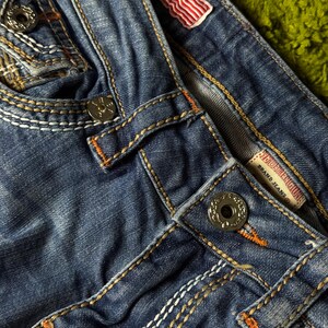 May include: Close-up of blue denim jeans with white and orange stitching. Features include metal buttons, a small American flag patch, and a brand label. The jeans have a worn, distressed look.